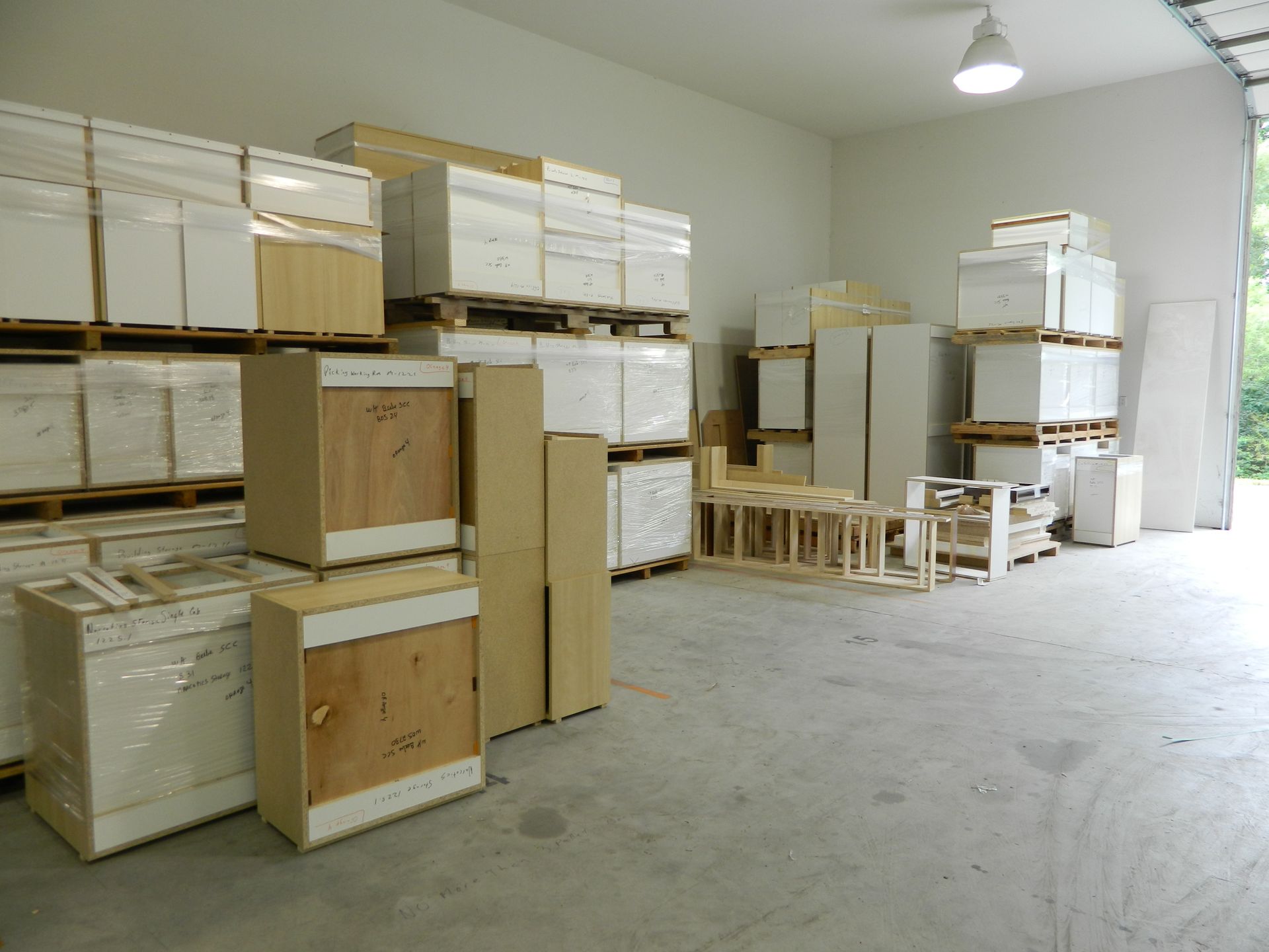 A warehouse interior with stacked boxes on pallets and assembled cabinets. An open door reveals greenery outside.