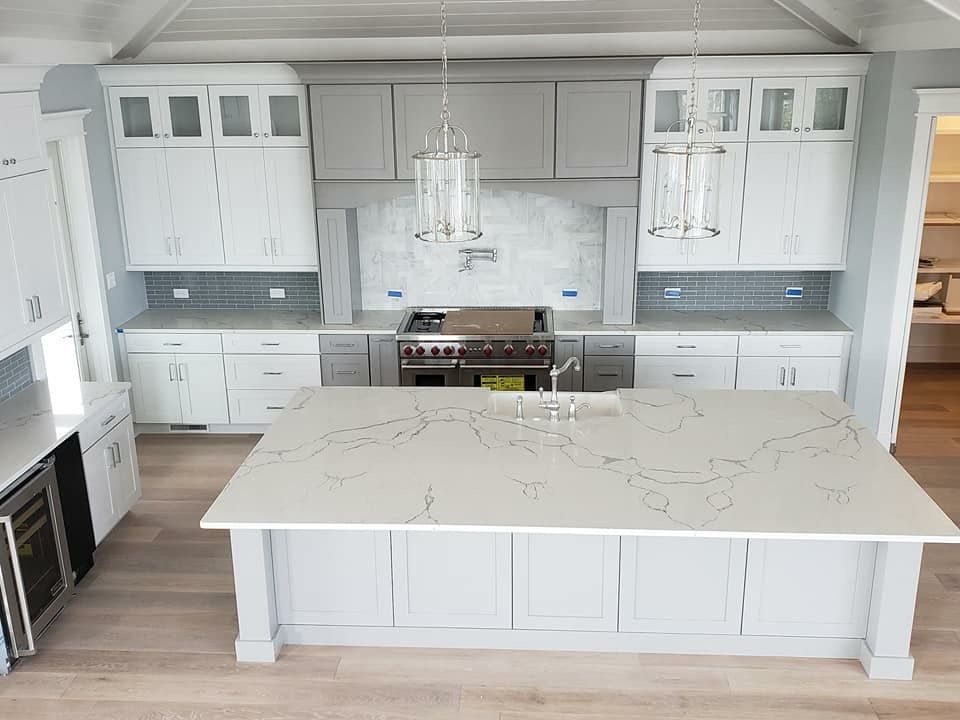 A modern kitchen with white and gray cabinets, a large island, and a stainless steel range. Light-colored quartz countertops and wood floors.
