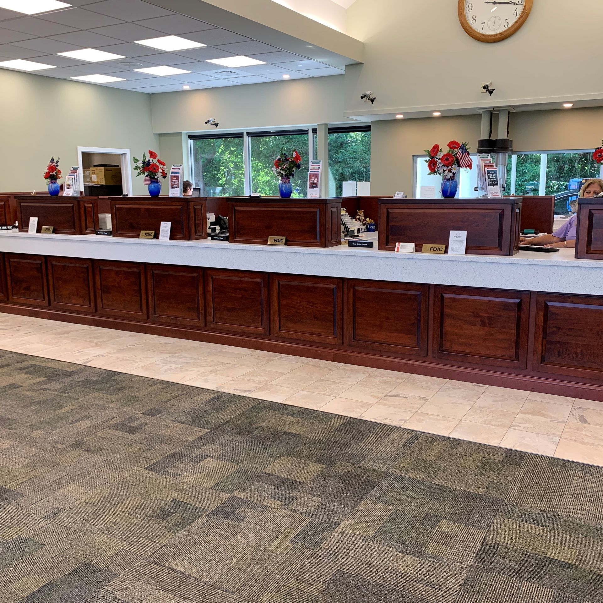 Bank teller counters with floral arrangements, a clock, and a window overlooking greenery.