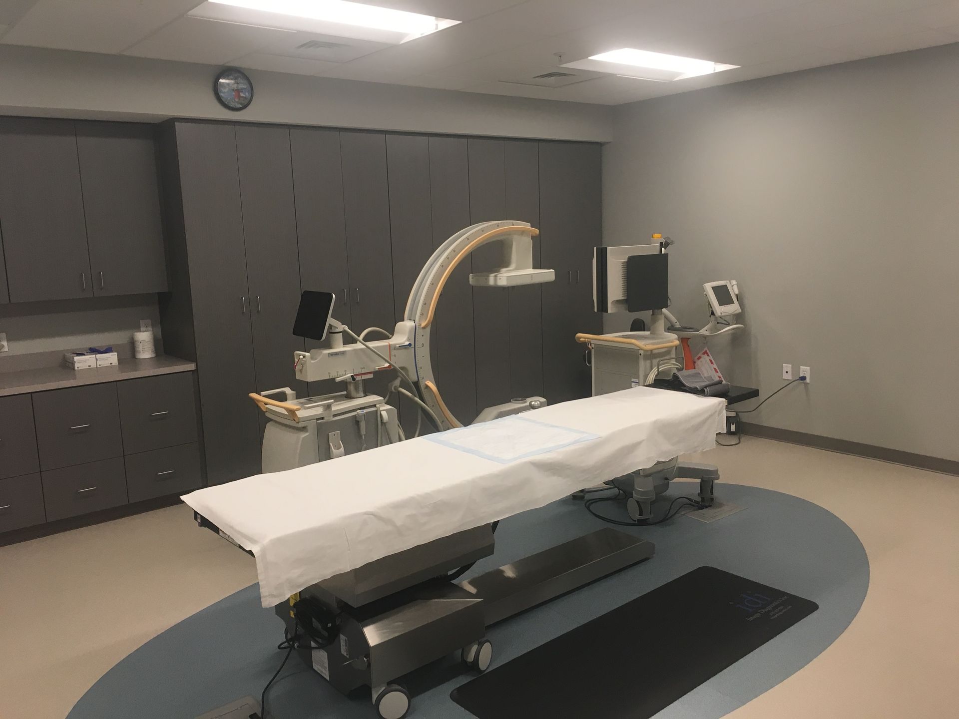 Medical room with an examination table covered in white, a C-arm machine, and medical monitors. Gray and blue floors.