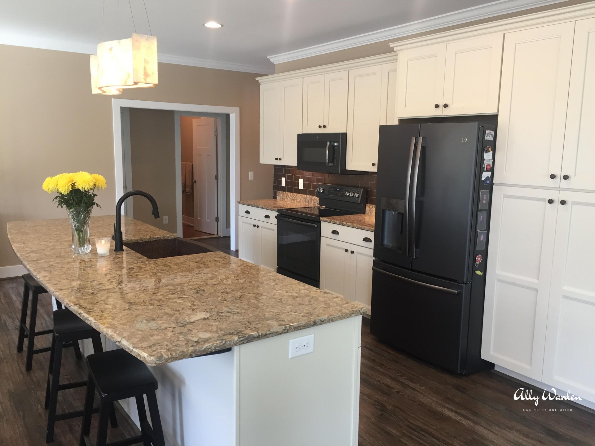 A modern kitchen with a large island and black appliances. Beige cabinets, granite countertops, and dark wood flooring.