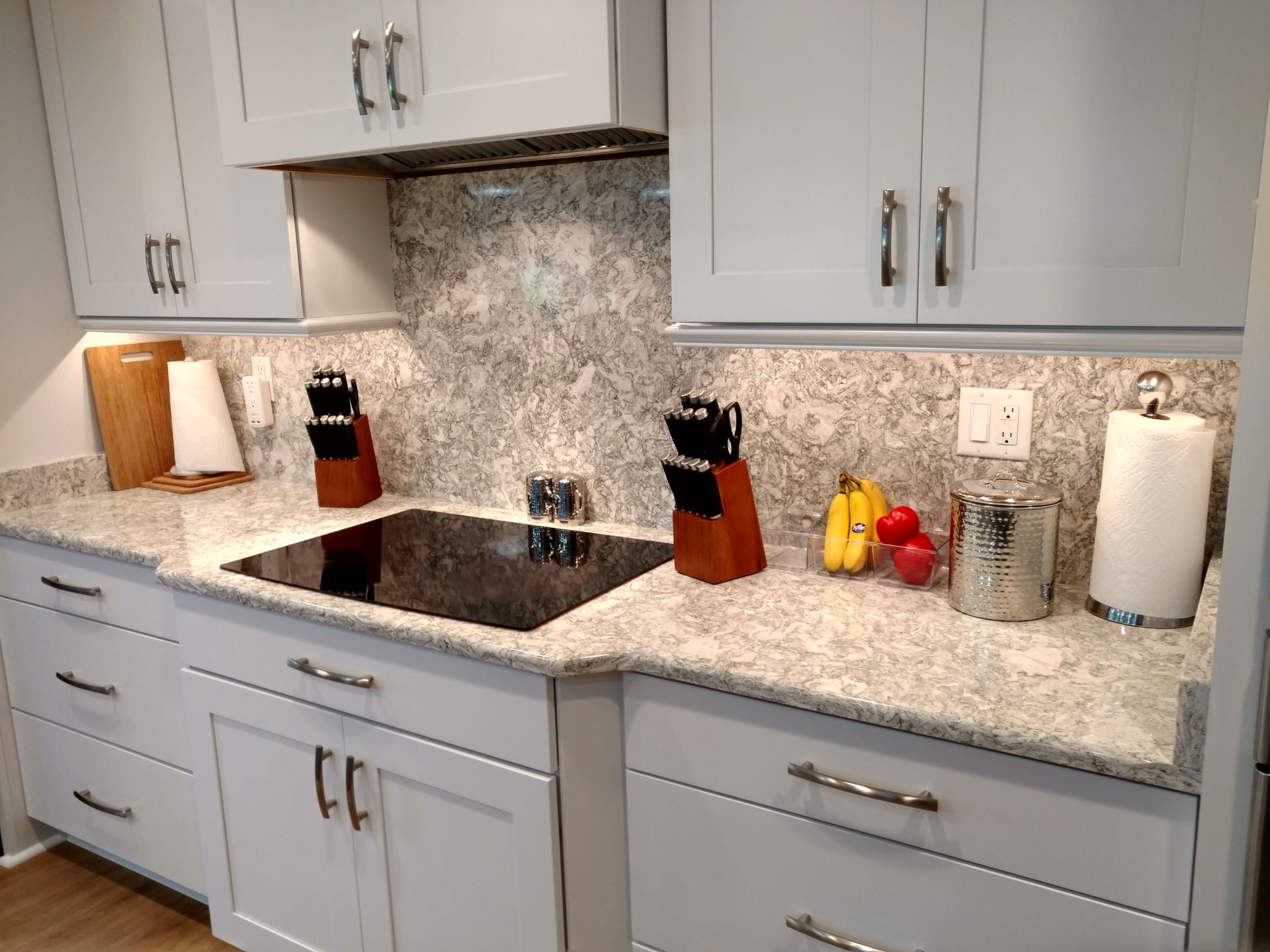 Light gray kitchen with white countertops, a black stovetop, and gray and white backsplash. Cabinets above and below.