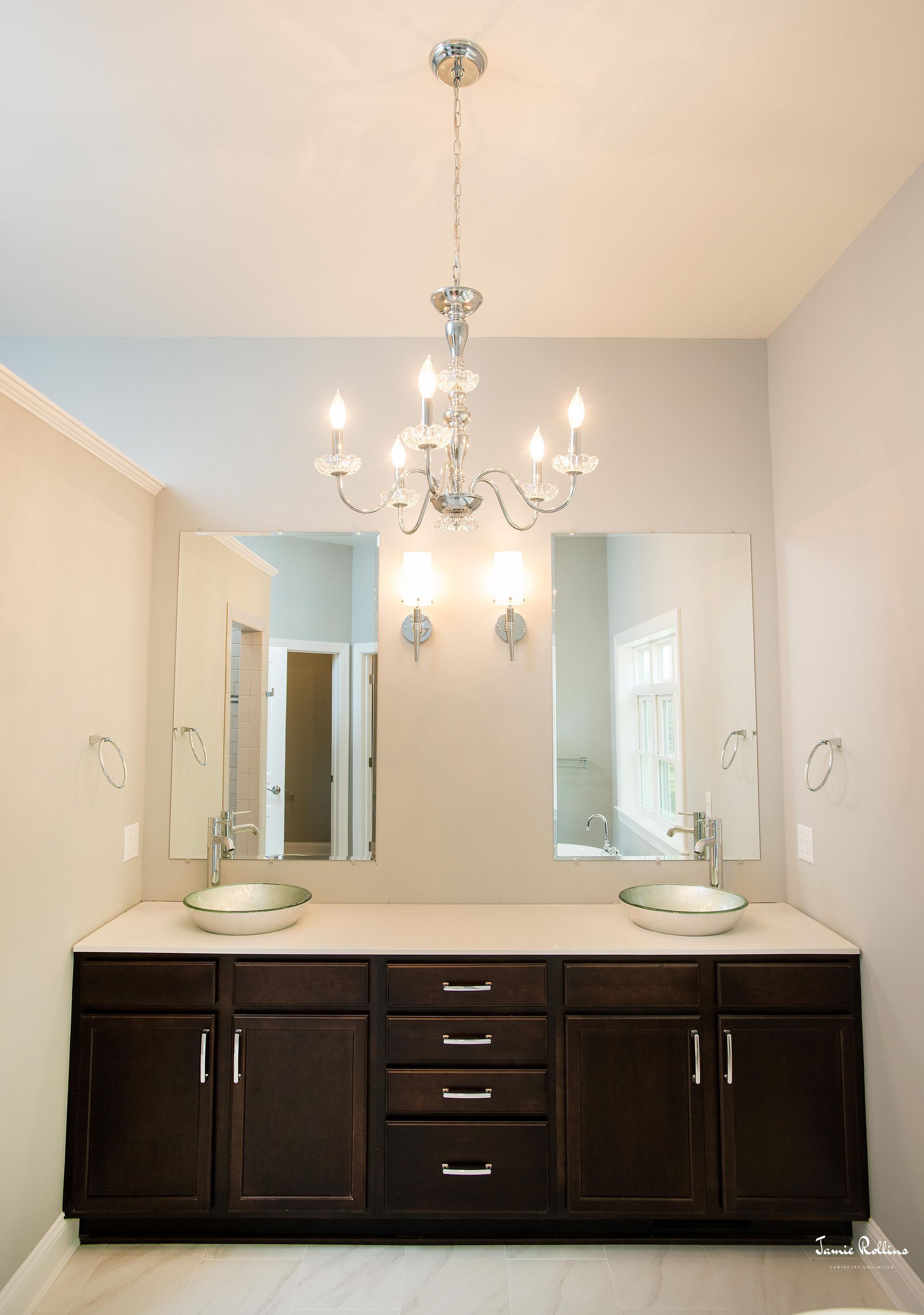 Bathroom with two sinks, dark brown cabinets, and a crystal chandelier. Two large mirrors reflect the room and a light gray wall.