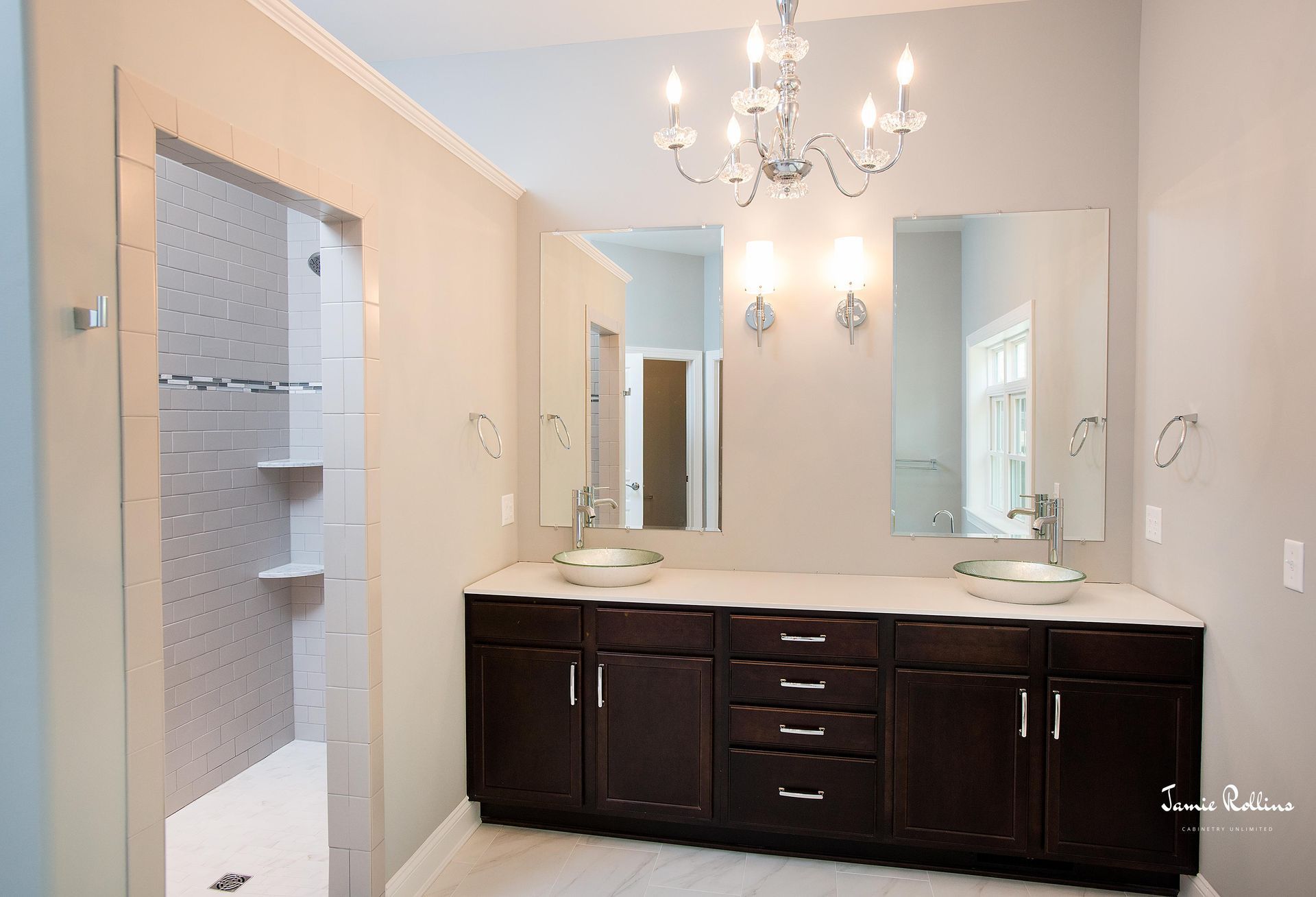 Bathroom with double sinks, dark brown cabinets, and a walk-in shower on the left. Two large mirrors above sinks reflect a hallway.