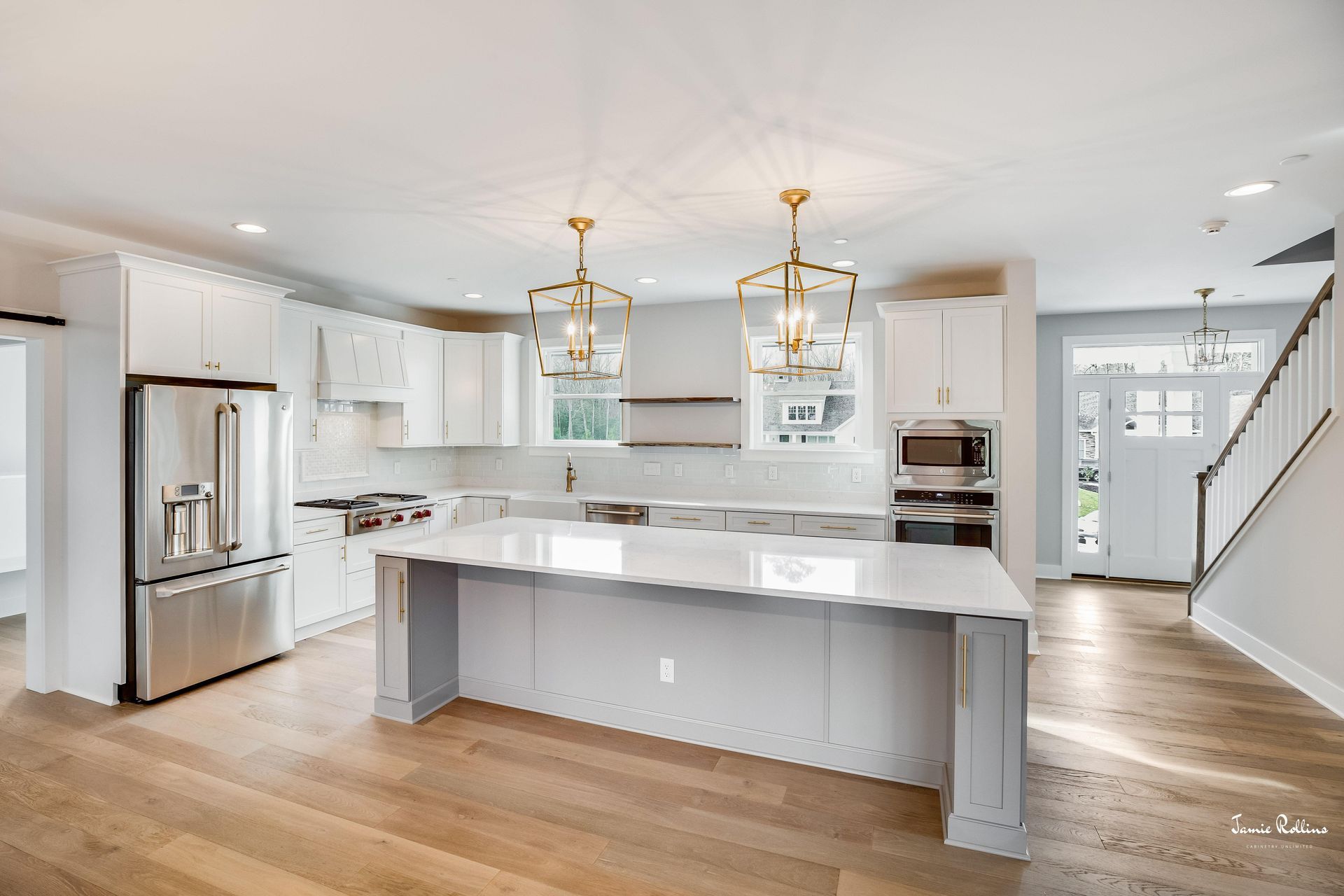 Modern white kitchen with a large island, stainless steel appliances, and gold pendant lights. Wooden floors lead to an entryway with stairs.