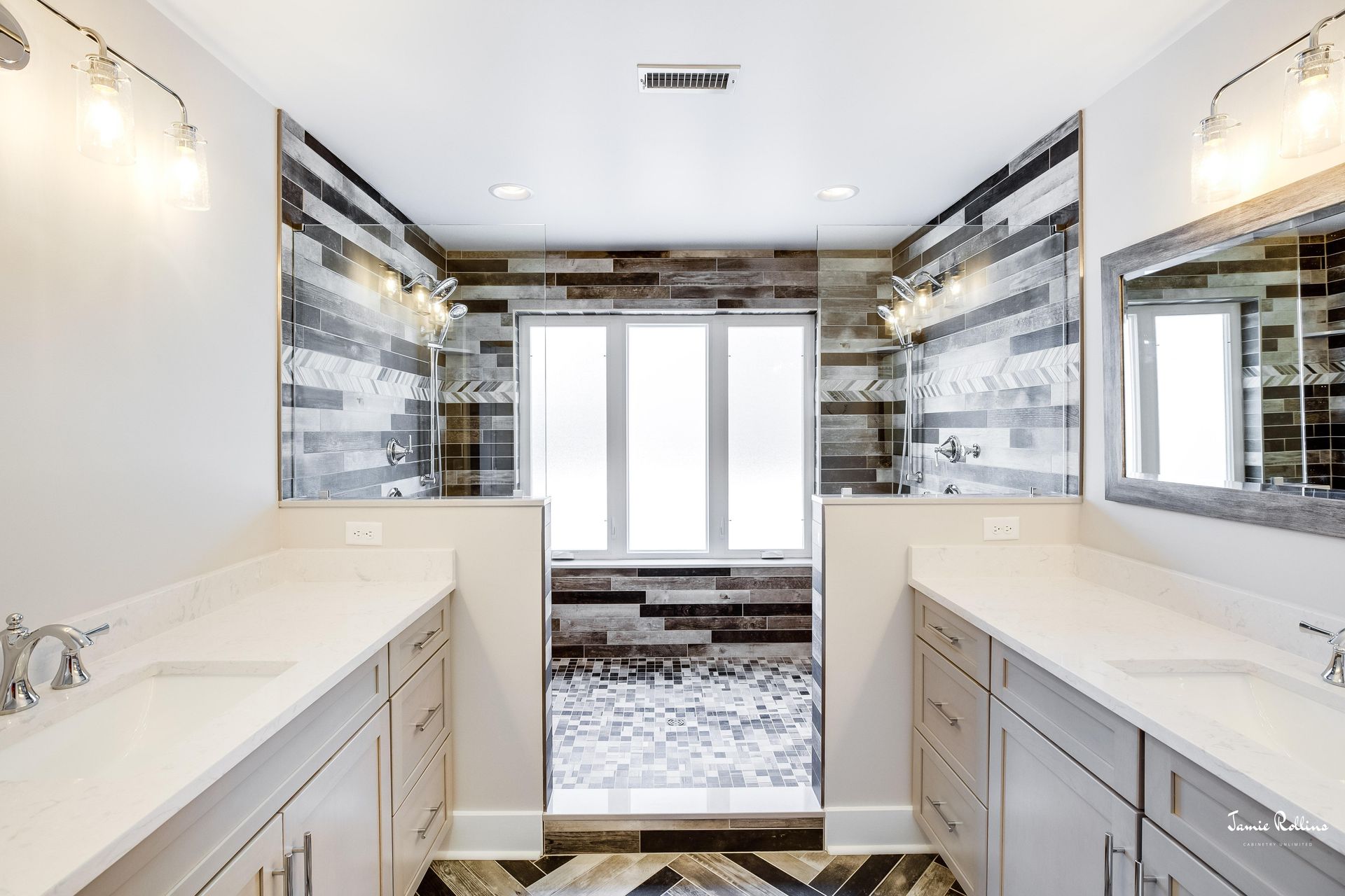 Spacious bathroom with dual vanities, glass-walled shower, and decorative tile. Light walls and cabinets with a geometric floor design.
