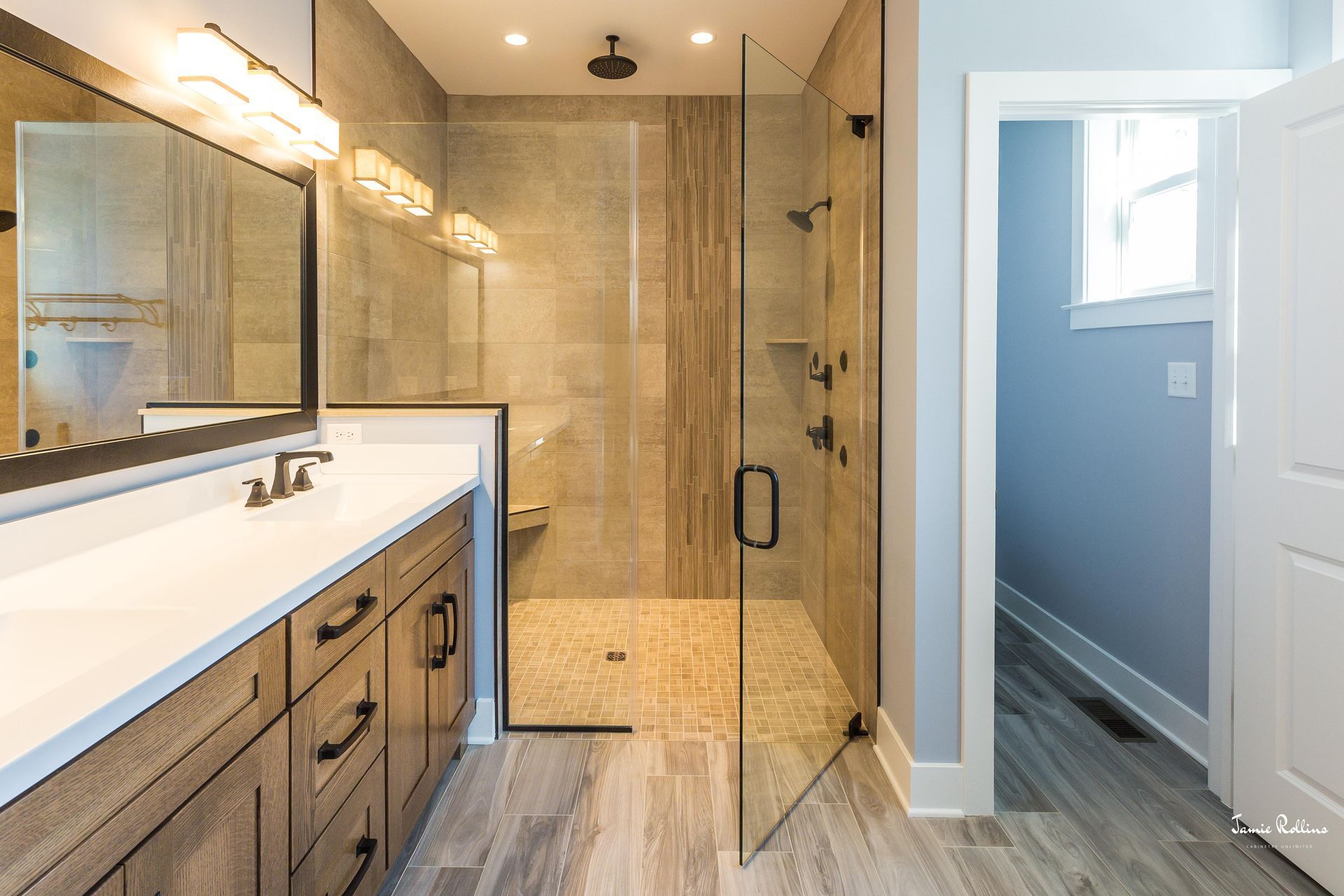 Modern bathroom with a walk-in shower, vanity, and an open doorway to another room. Neutral tones, wood cabinets, and a glass shower door.