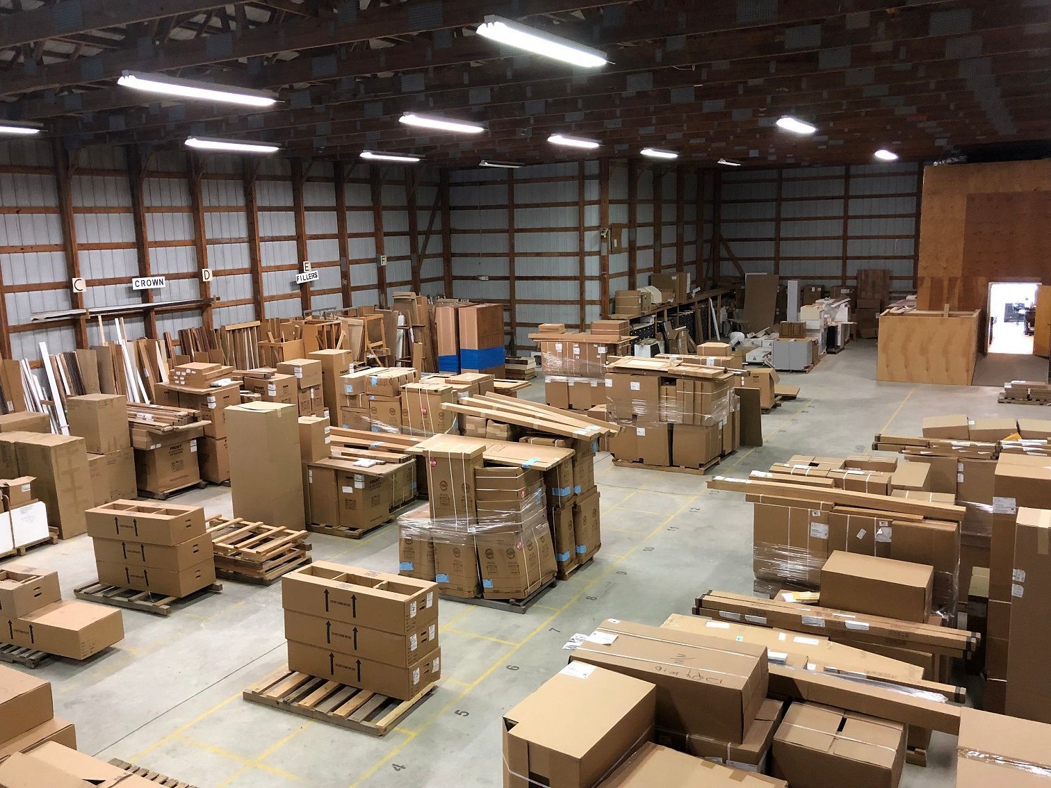 Warehouse interior with cardboard boxes, some on pallets, stacked throughout the space. Large overhead lights and metal framework visible.