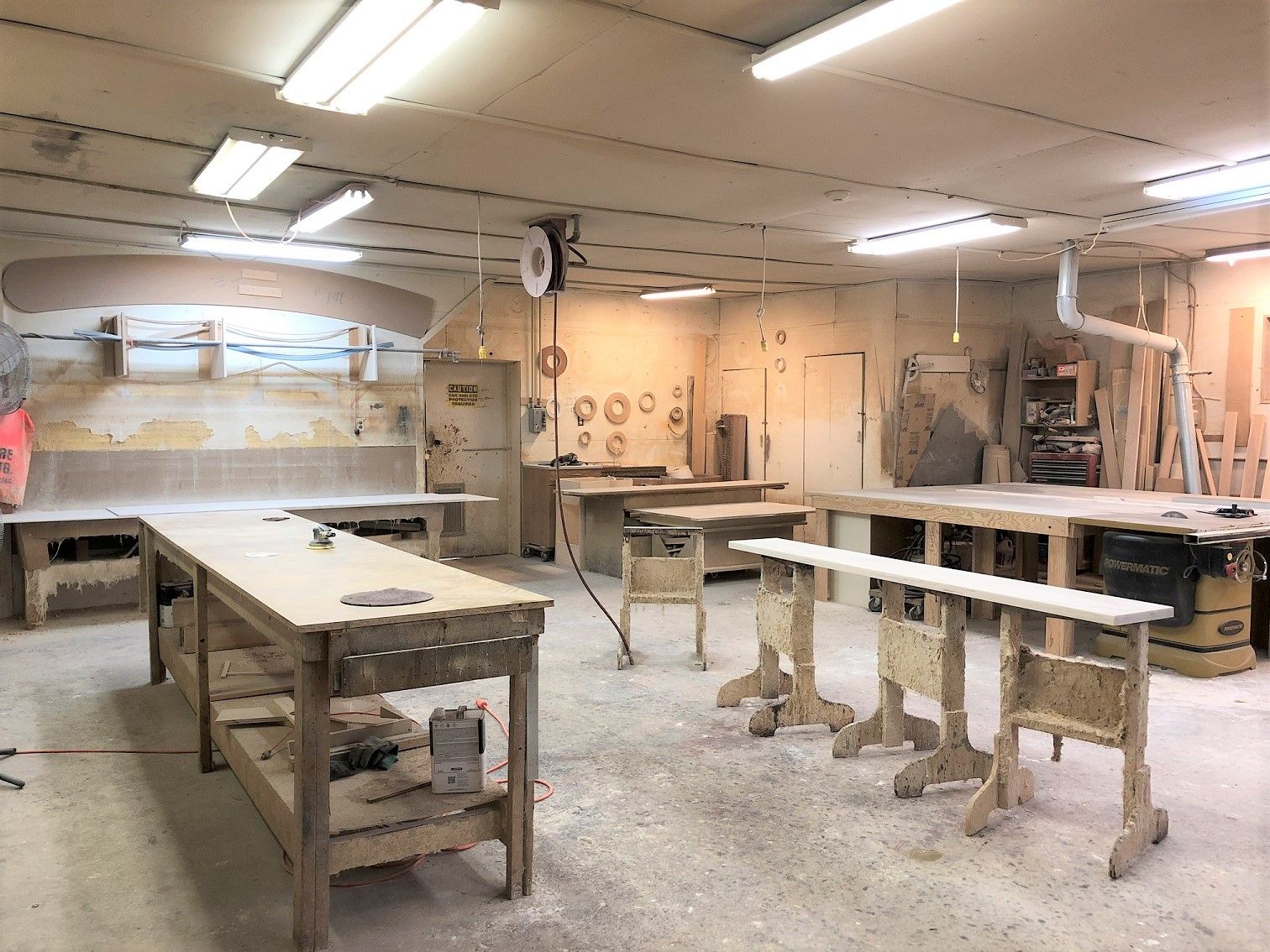 A dusty workshop with multiple wooden workbenches and tools. The room is lit by fluorescent lights, with wood scraps on the floor.