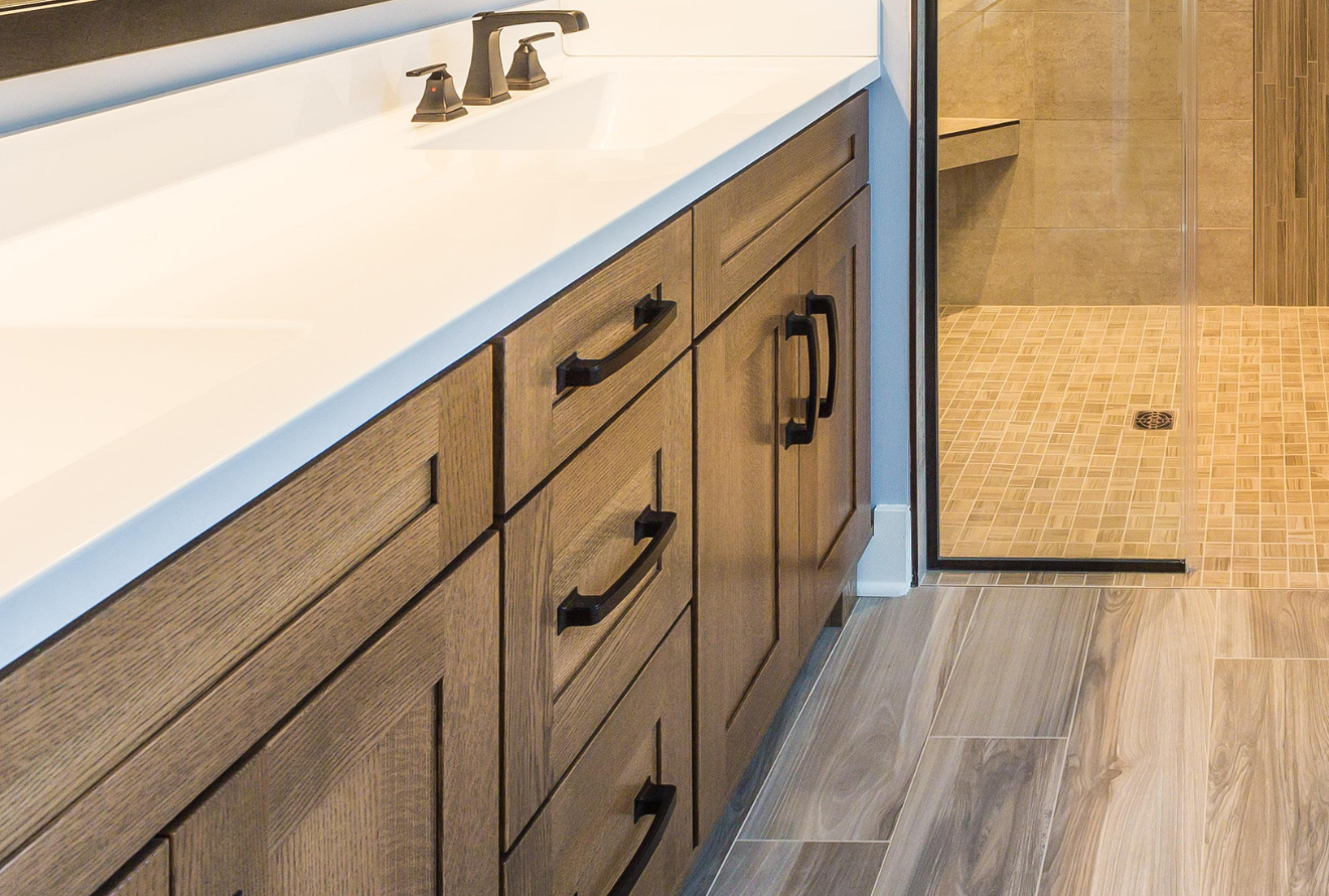 Bathroom vanity with a white countertop, wooden cabinets, and black hardware. A glass shower is visible in the background.