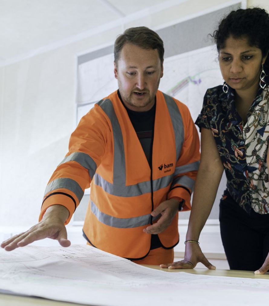 Man in orange safety vest points at a blueprint with a woman, inside a brightly lit office with maps on the wall.