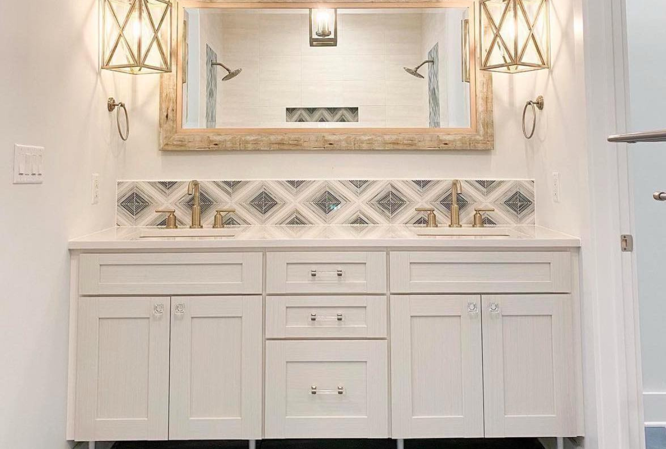 Bathroom with a white vanity, patterned tile backsplash, and a large, wooden-framed mirror. Gold light fixtures and faucets add accents.