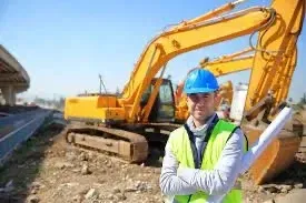 A construction worker is standing in front of a yellow excavator on a construction site.
