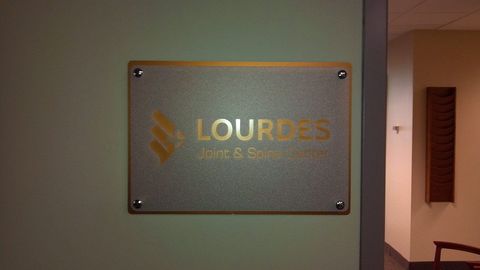 Sign for Lourdes Joint & Spine Center, gold text and logo on frosted background, mounted on a wall.