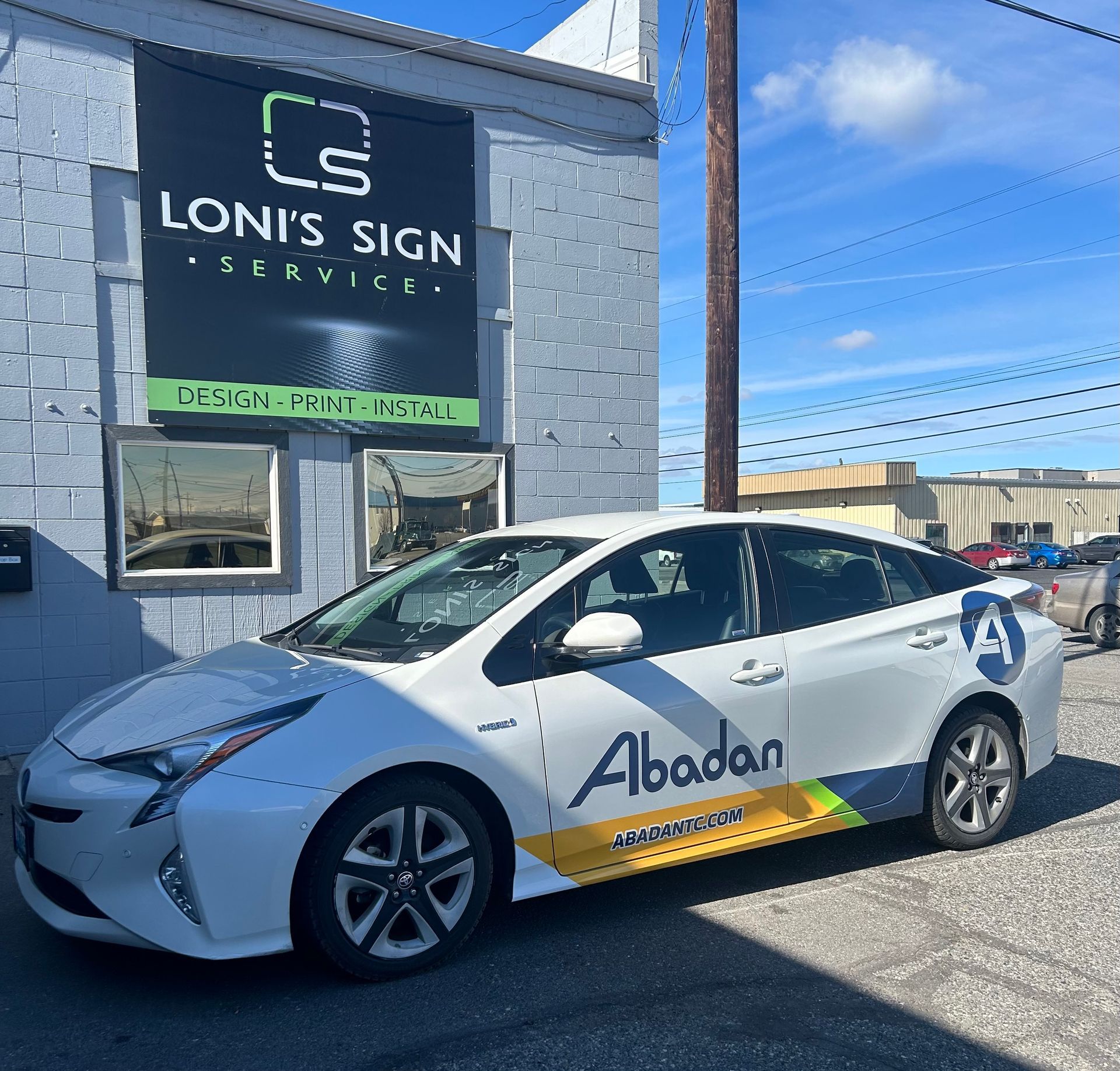 White car with Abadan logo in front of Loni's Sign Service building on a sunny day.