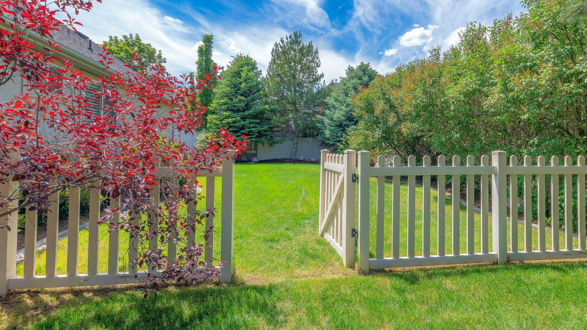 Open white gate in a green lawn, framed by red and green trees under a blue sky.