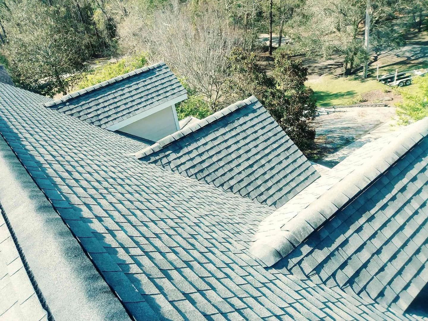 View of a multi-gabled roof with gray shingles against a blurred outdoor backdrop of trees.