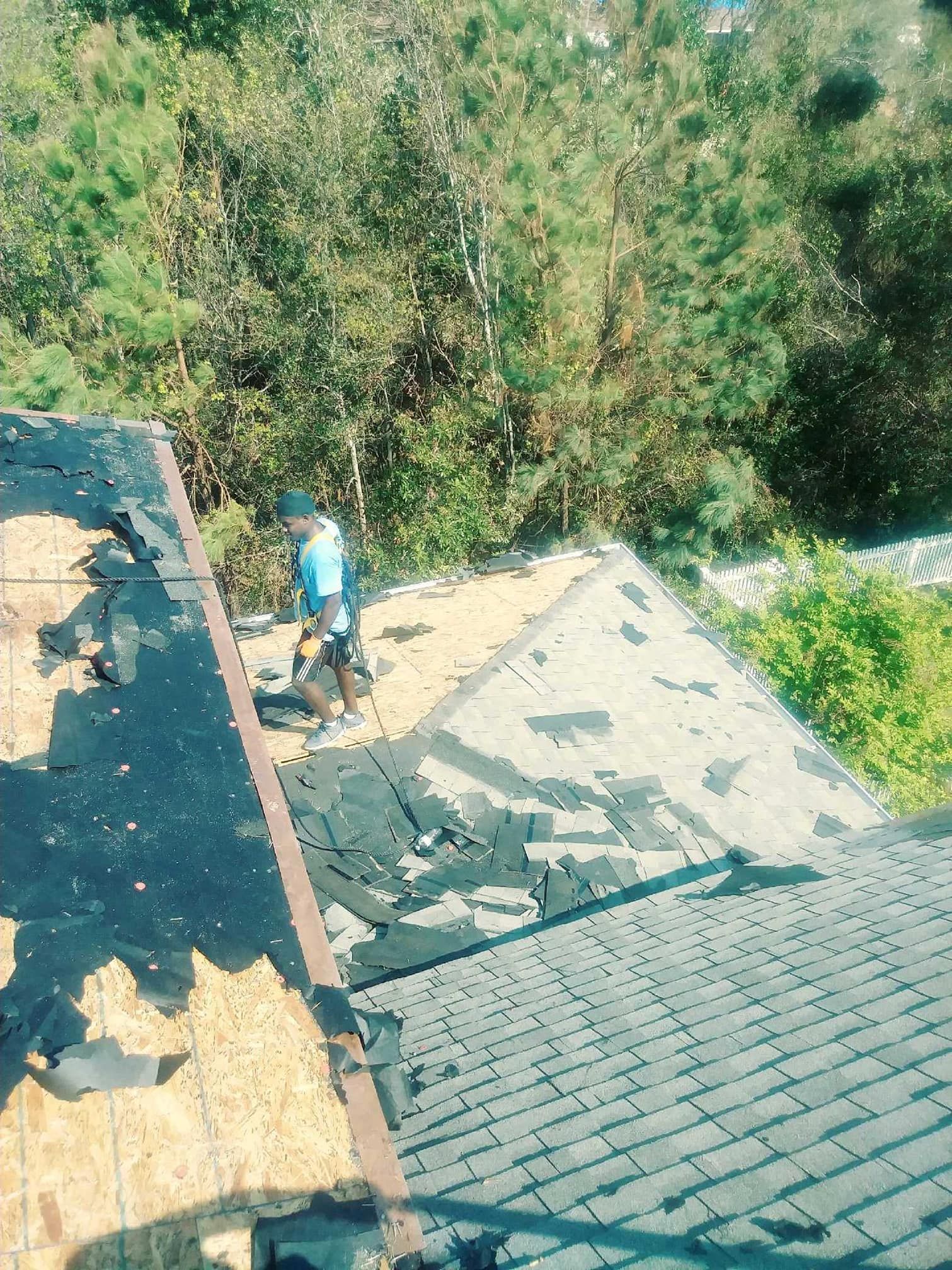 Man removing shingles from a rooftop with trees in the background on a sunny day.