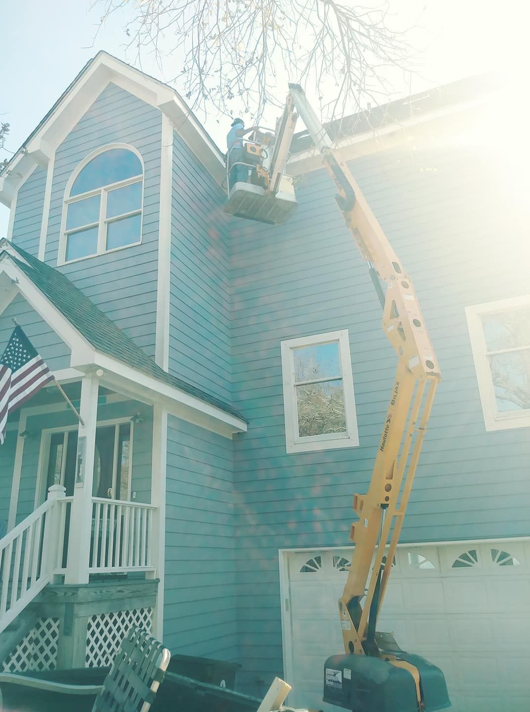 Person in a lift working on the side of a blue house with an American flag, sunny day.