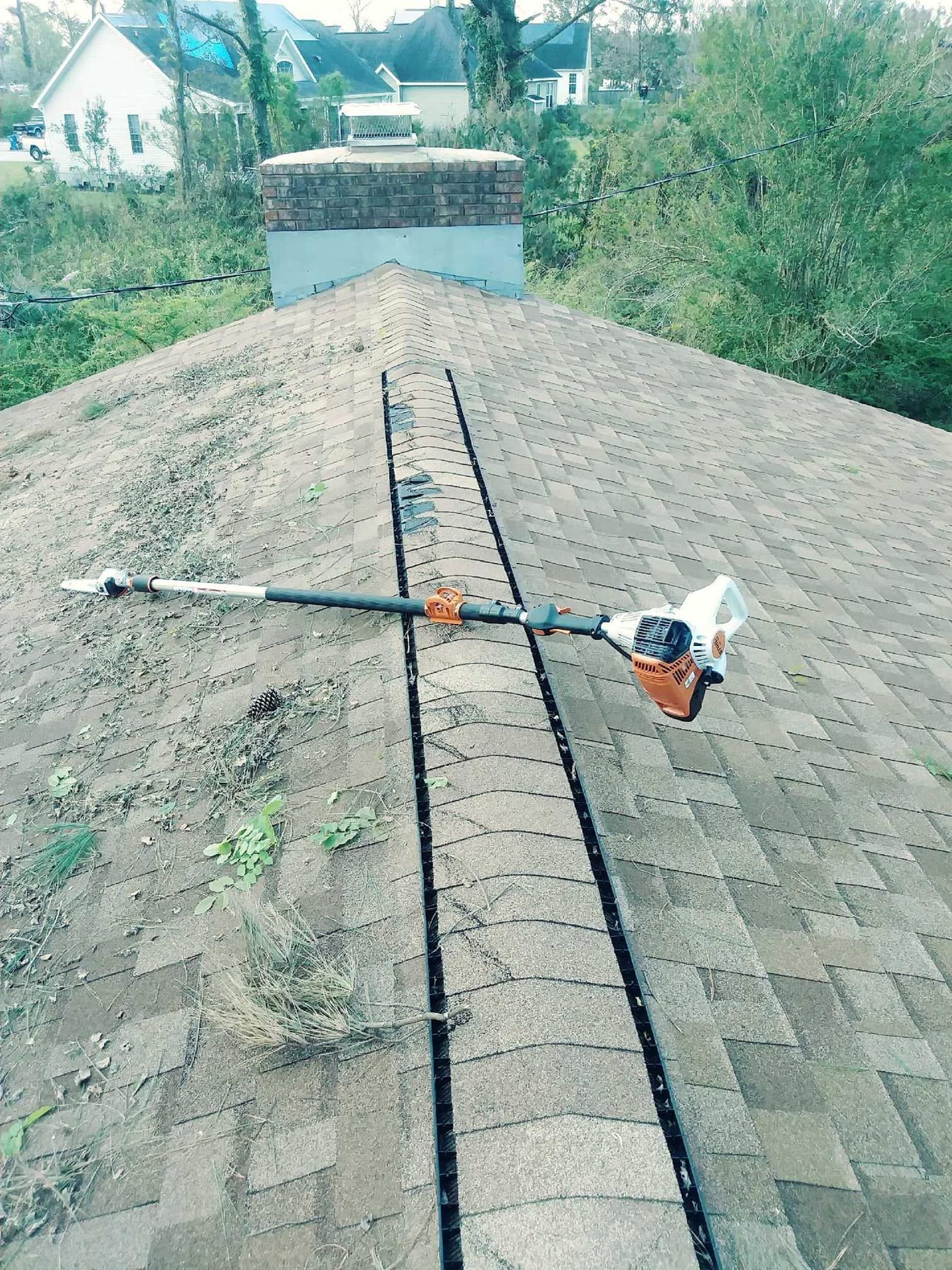A person uses a pole saw on a rooftop to trim the area along the roof's vent.