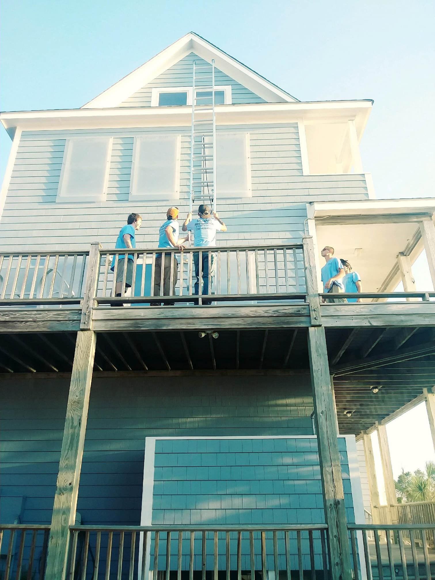 People on a house deck preparing for a storm, boarding up windows. Blue house, sunny day.