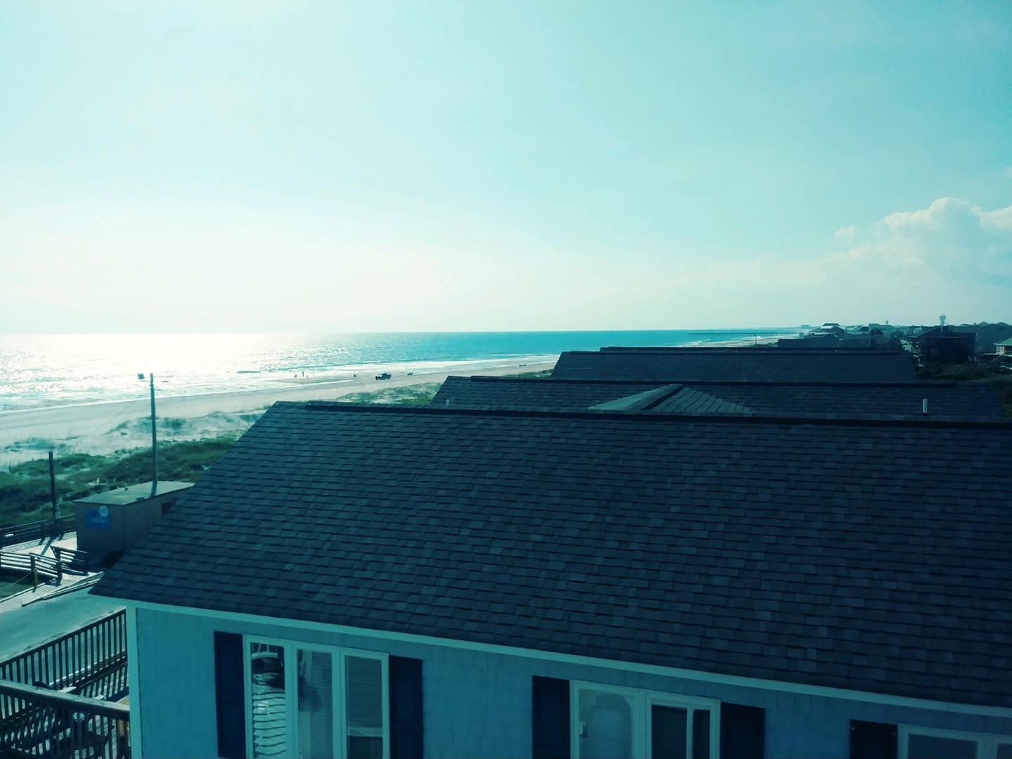 Ocean view from a house roof, blue water, sky, and sunlight with beachfront houses in the background.