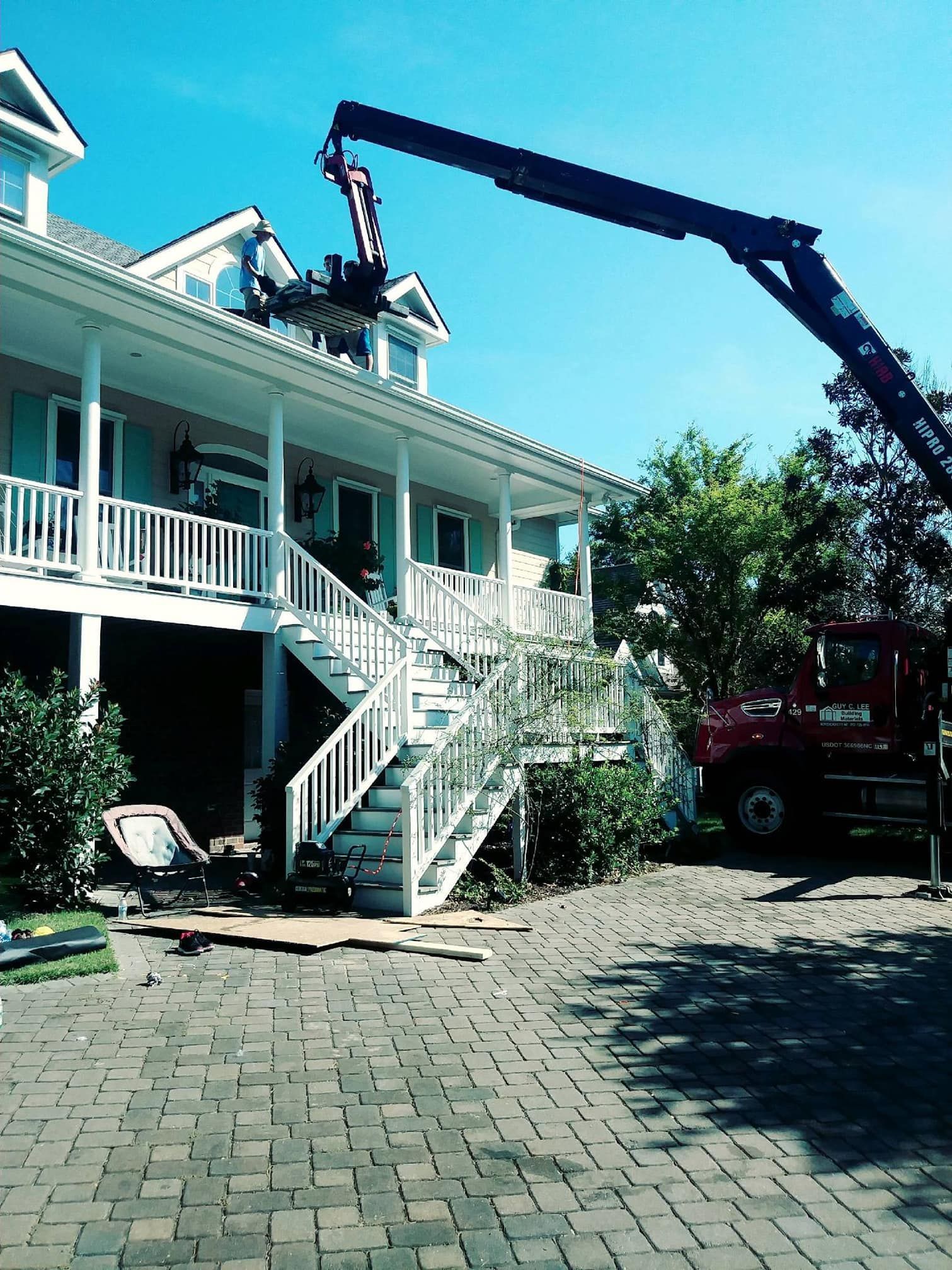 Crane lifting objects to the roof of a house with a white porch and stairs. A red truck is nearby.