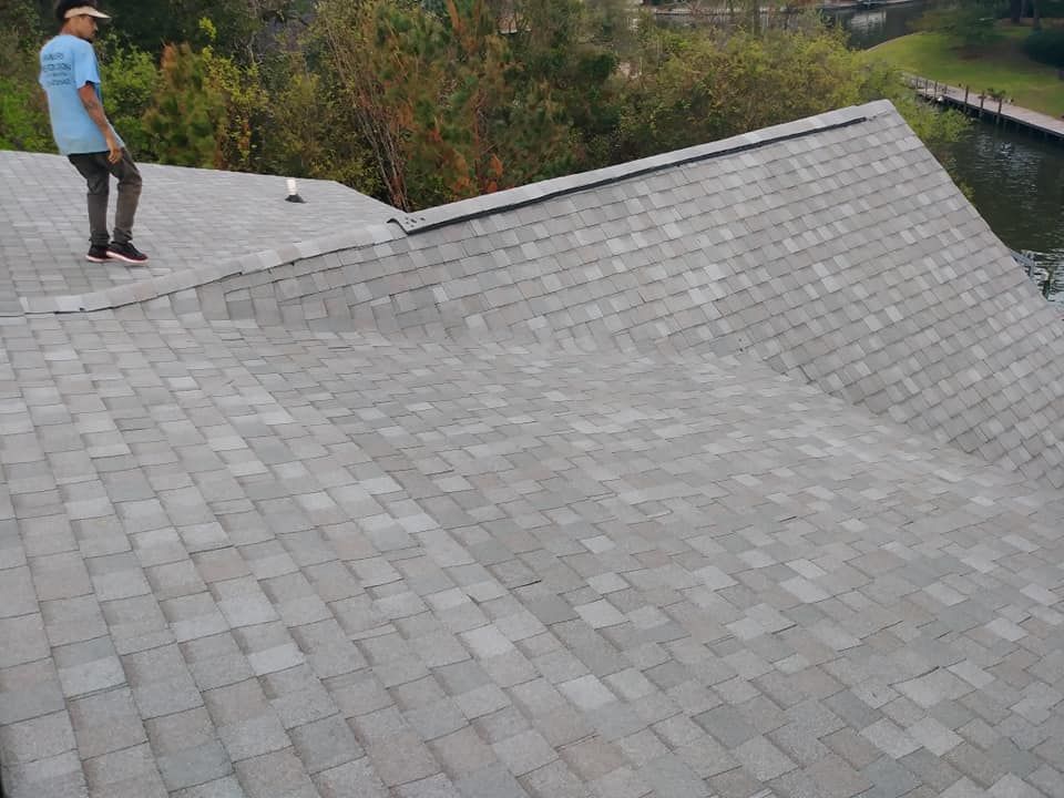 Man on a gray shingled roof. He is near a lake.