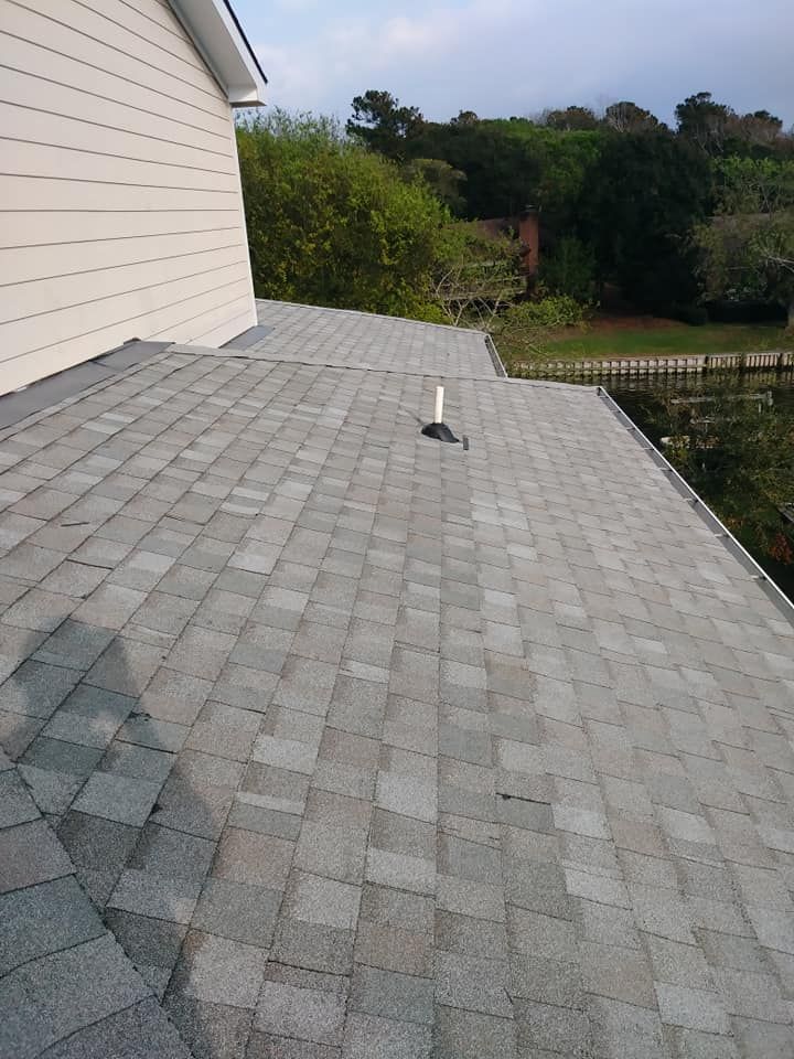 Gray shingle roof with a vent pipe, attached to a light beige house, with trees and fence in the background.
