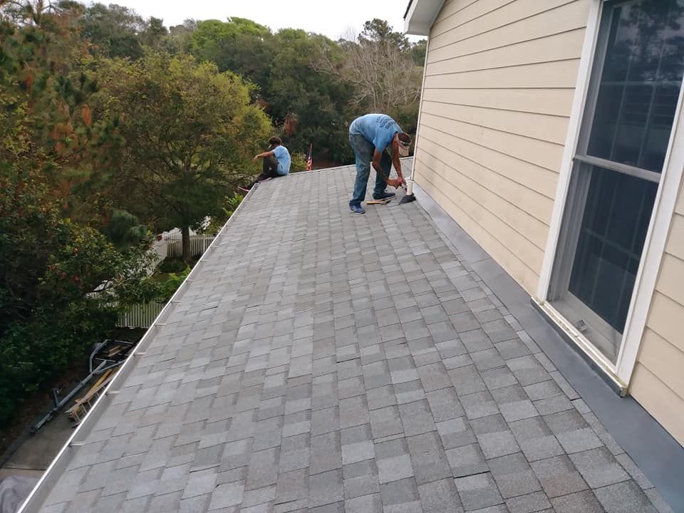 Workers on a roof repairing shingles, near a building and trees.