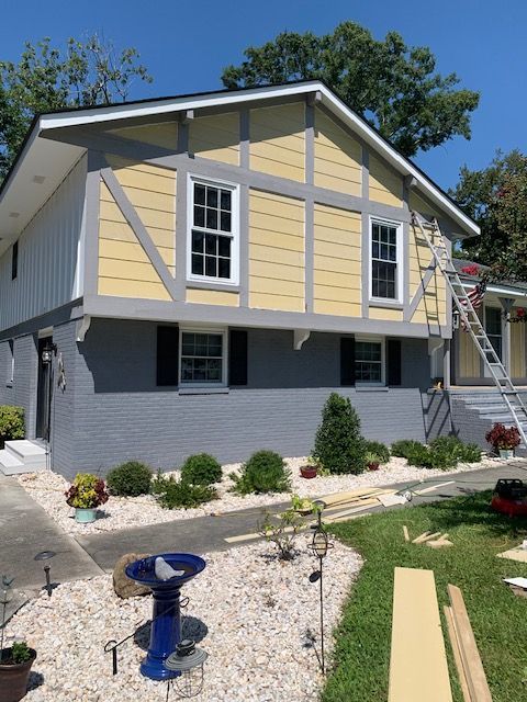 Two-story house with yellow and gray facade; worker on ladder. Landscaping and blue birdbath in front.
