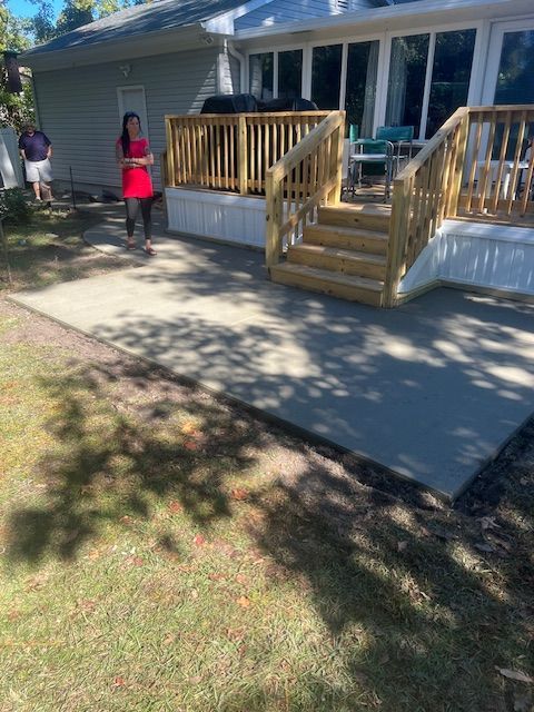 Person stands on a new concrete patio leading to a wooden deck with stairs. The background shows a house.