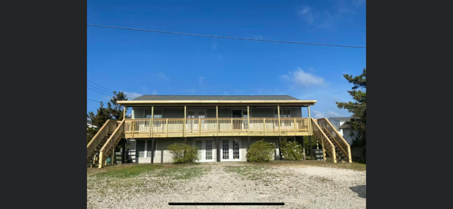 Two-story beach house with wooden decks, stairs, and a gravel yard under a blue sky.