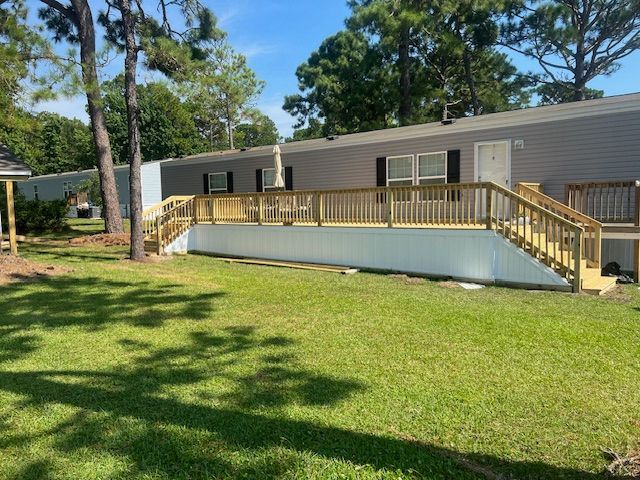 A gray mobile home with a wooden deck, on a green lawn, in sunlight.