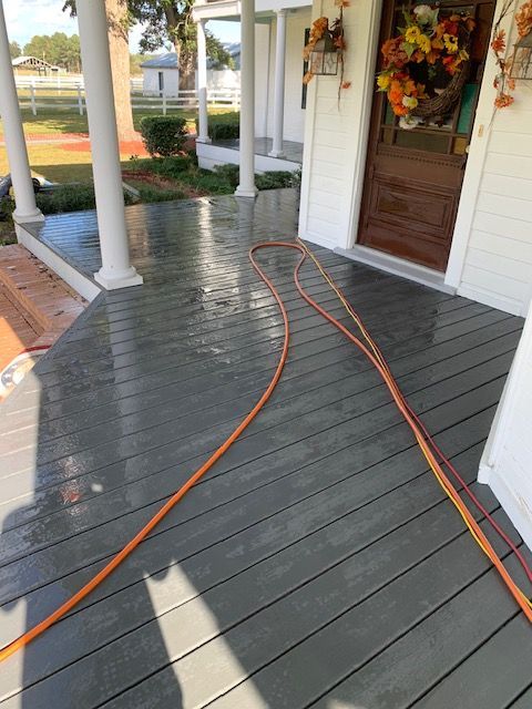 A porch with a wet, gray wooden deck, and an orange hose across it. A brown door and wreath are visible.