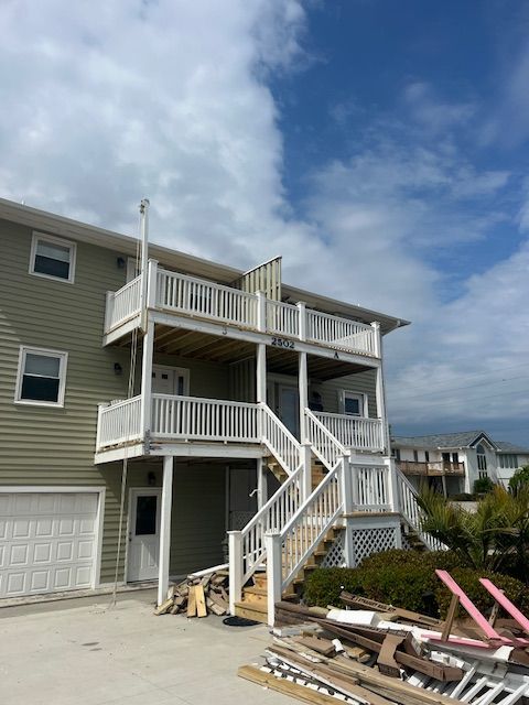 A two-story beach house with white railings, stairs, and an open balcony under a cloudy sky.