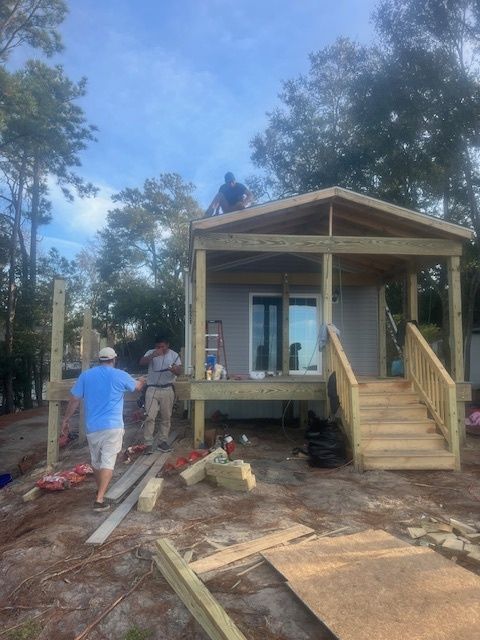 Construction workers building a small house with a porch and stairs outdoors on a sunny day.