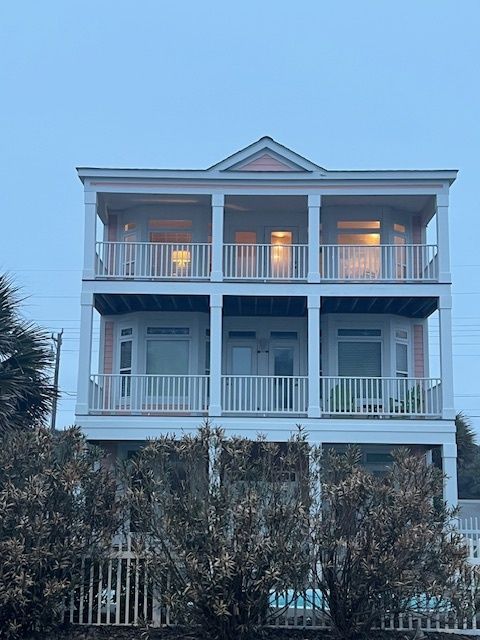 Three-story light pink house with white columns and balconies, viewed over bushes, under an overcast sky.