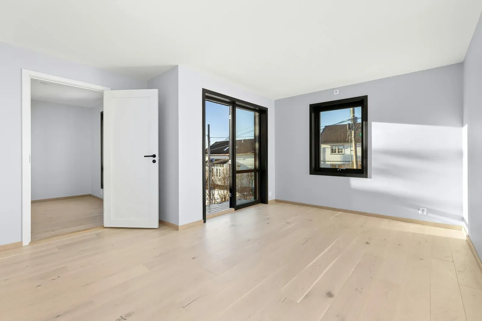 Empty, light-filled room with light wood floors, light blue walls, and windows/door leading outside.