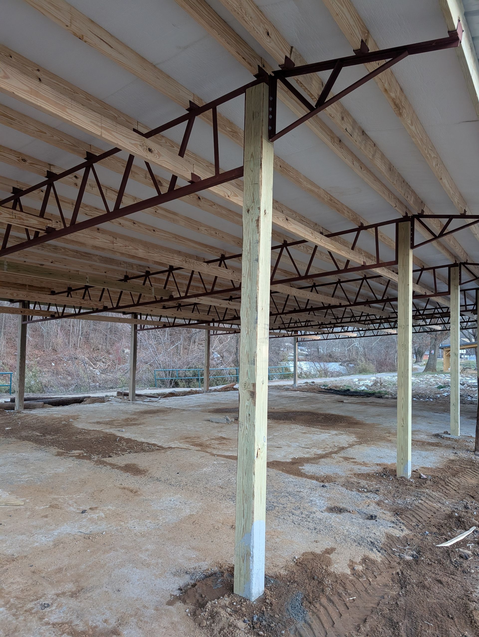 Open-air structure under construction. Tall concrete columns support a wooden ceiling and metal trusses, set in a dirt area.