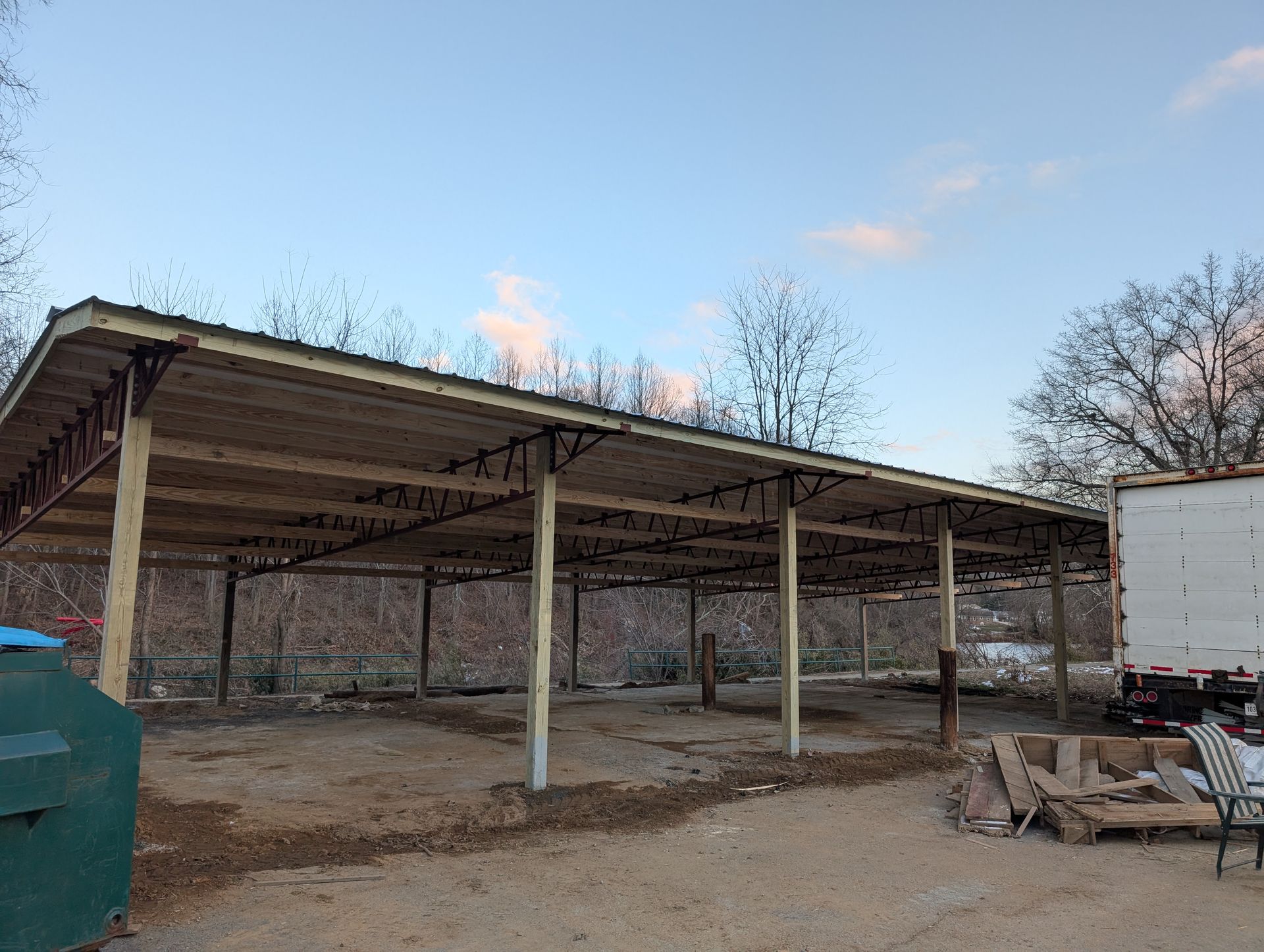 A large, unfinished wooden carport under a partly cloudy sky. It's set on a dirt and gravel surface with a few trees in the background.