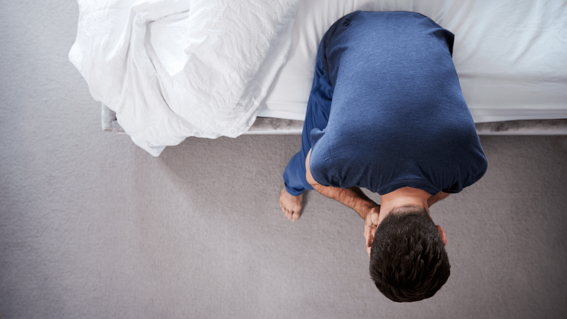 An overhead shot of a man sat on a bed, his head in his hands. He is wearing a blue shirt.