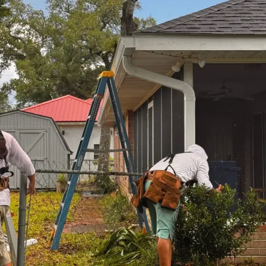Workers in white shirts and tool belts work on the exterior of a house near a blue ladder and a chain-link fence.
