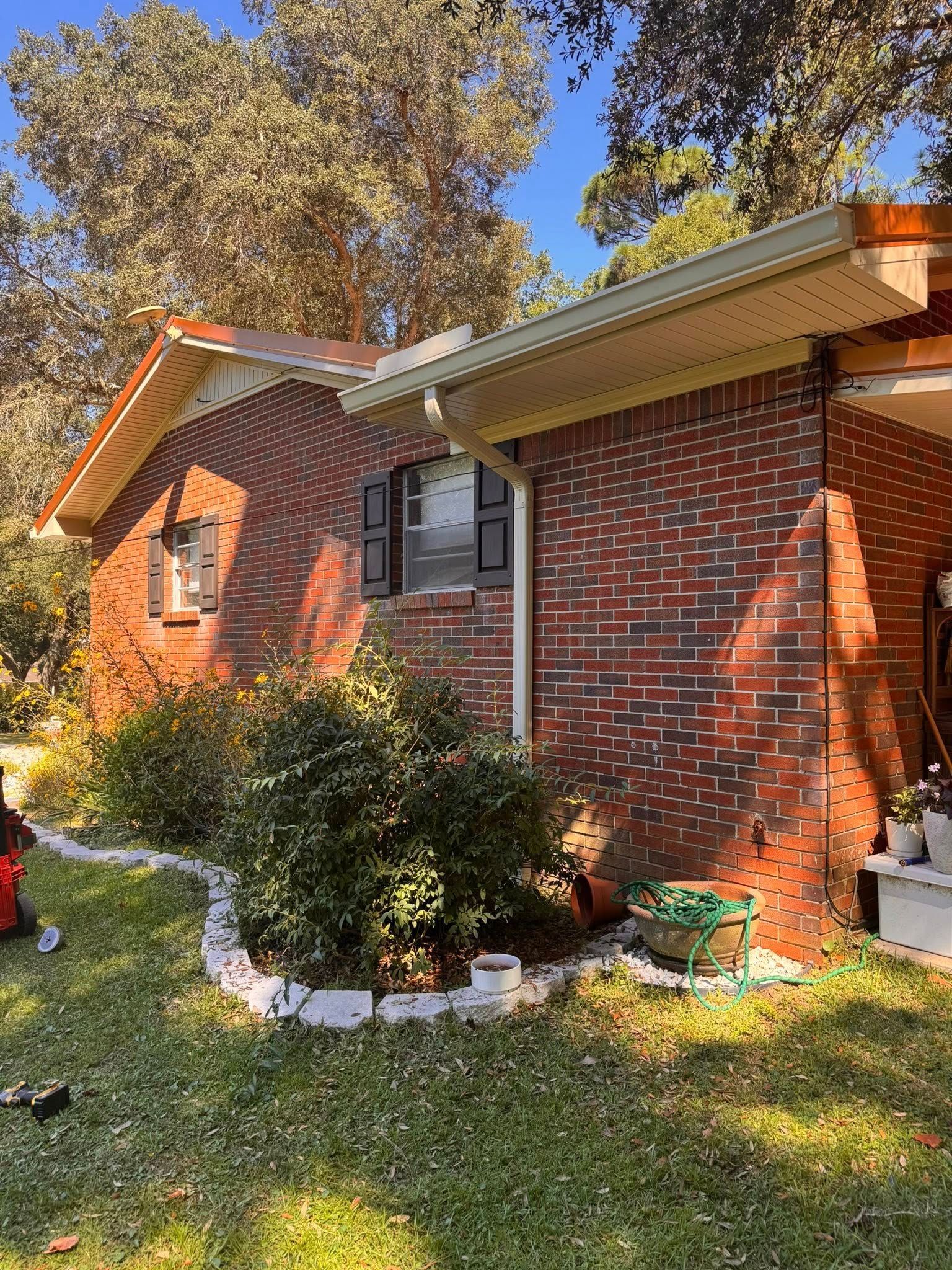 A red brick ranch-style house exterior with dark window shutters, a covered porch, and bushes in the front yard.