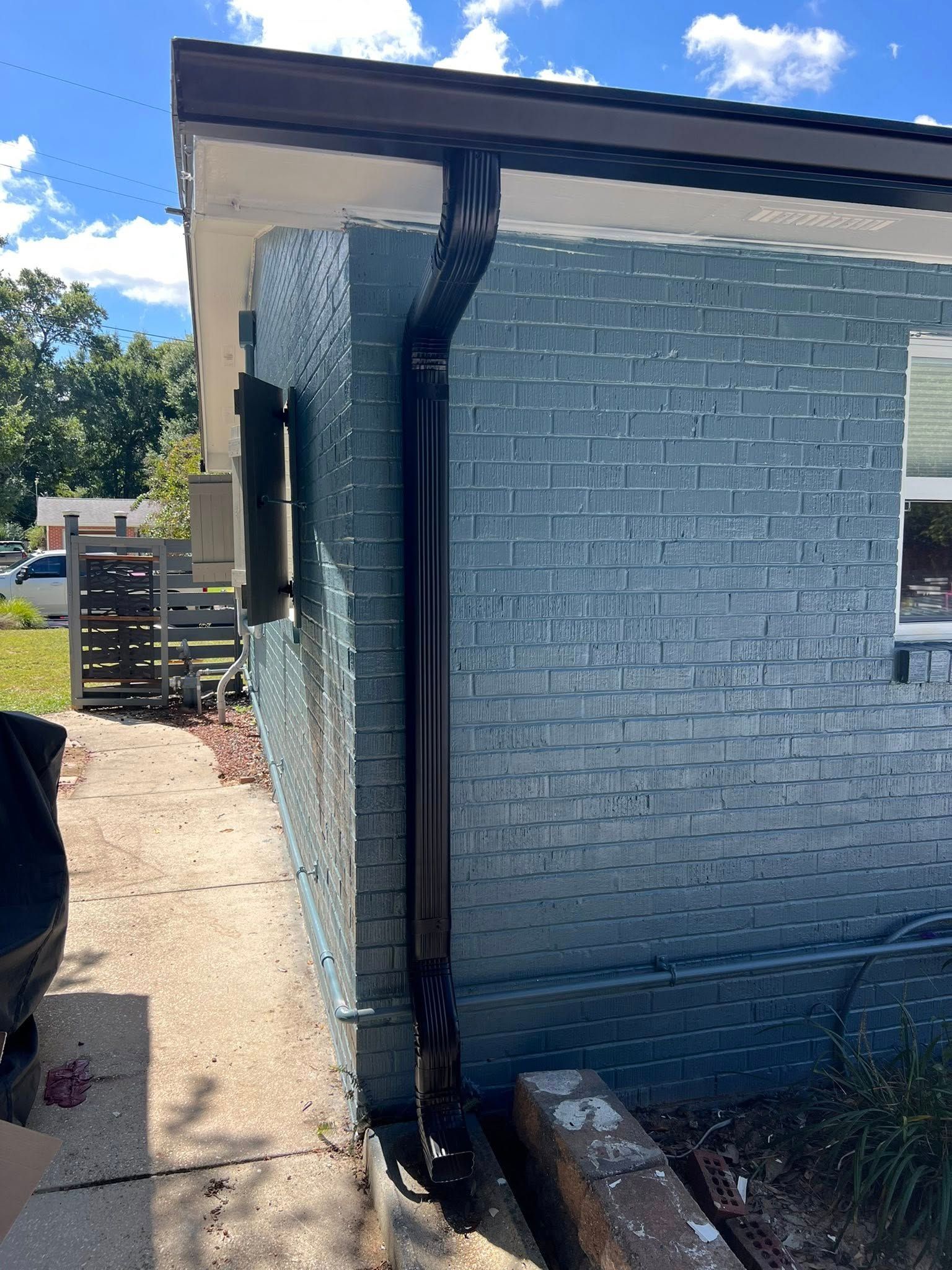 A black gutter downspout attached to the corner of a house with blue-painted brick walls, leading to a concrete drain.