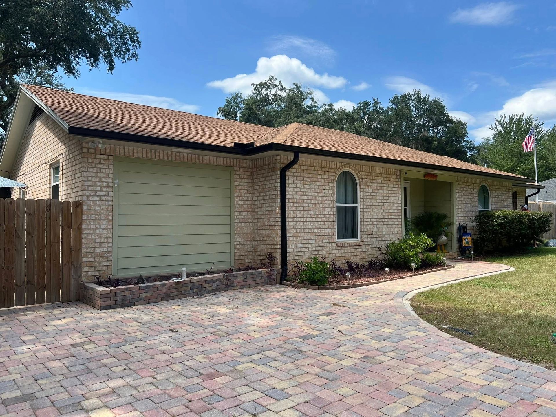 A single-story brick house with a paved driveway, light-green garage door, arched window, and a wooden fence.