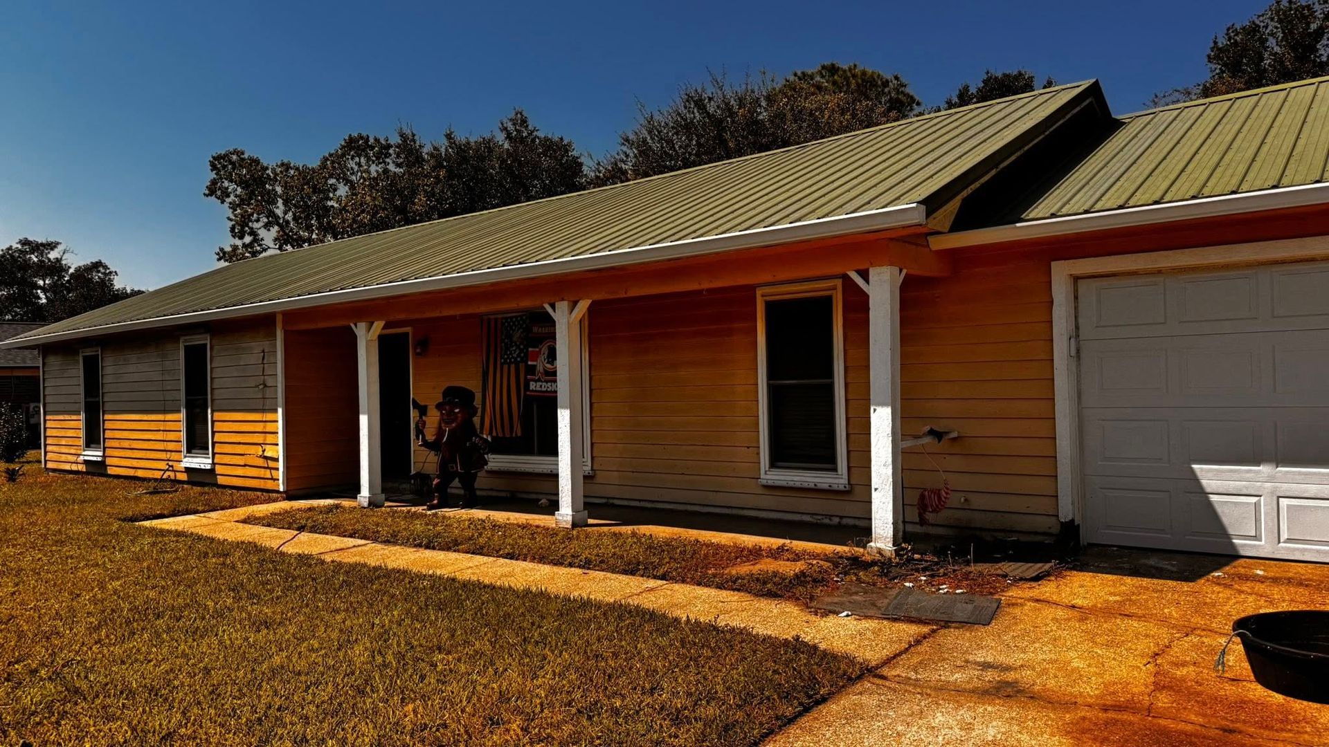 A one-story yellow house with a covered front porch and a green metal roof under a clear blue sky.