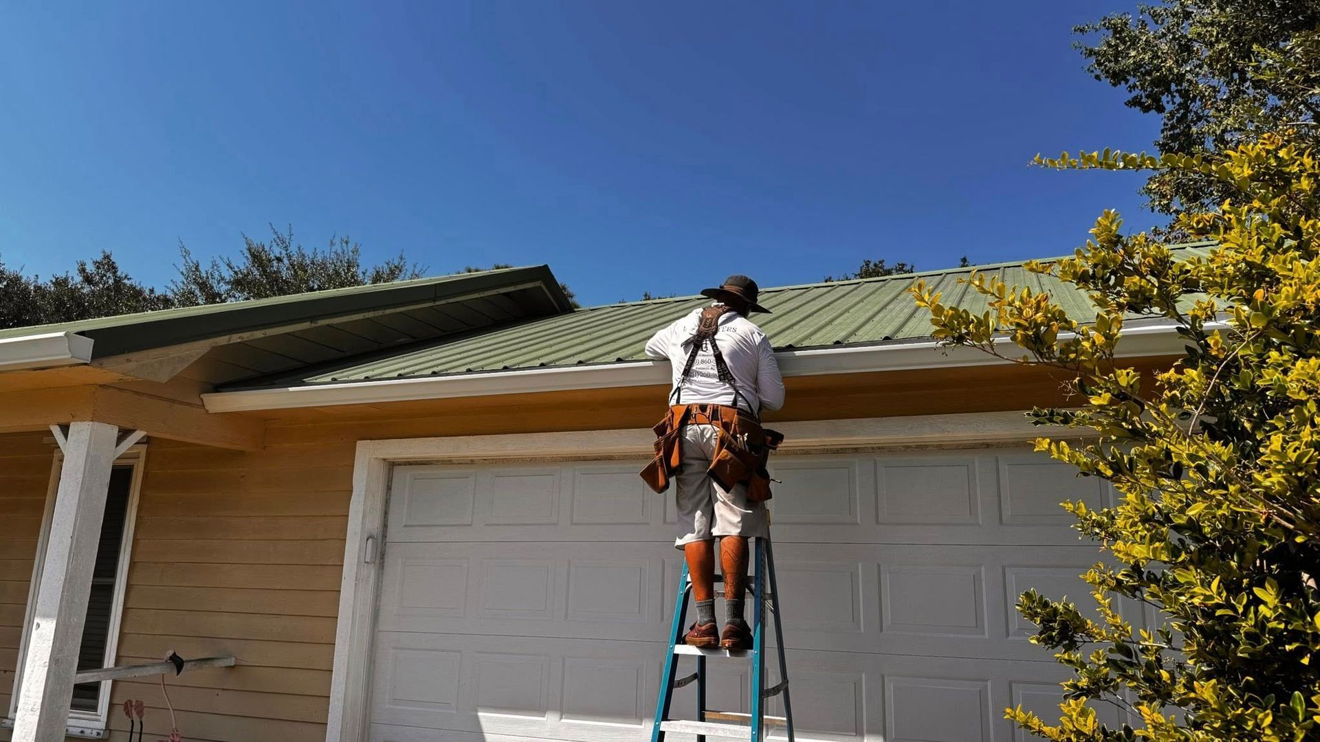 A person in a hat and tool belt stands on a ladder working on the roof of a house with a garage.