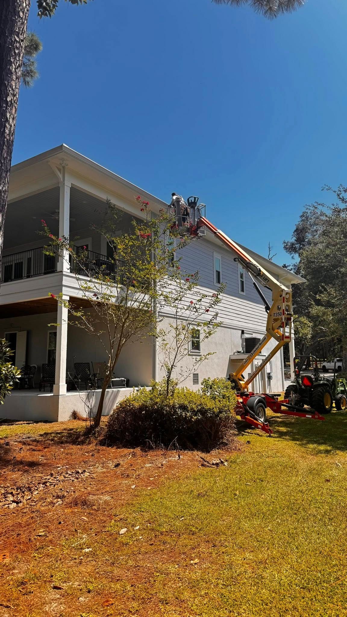 Workers in an aerial lift platform work on the roof of a two-story white house on a sunny day.