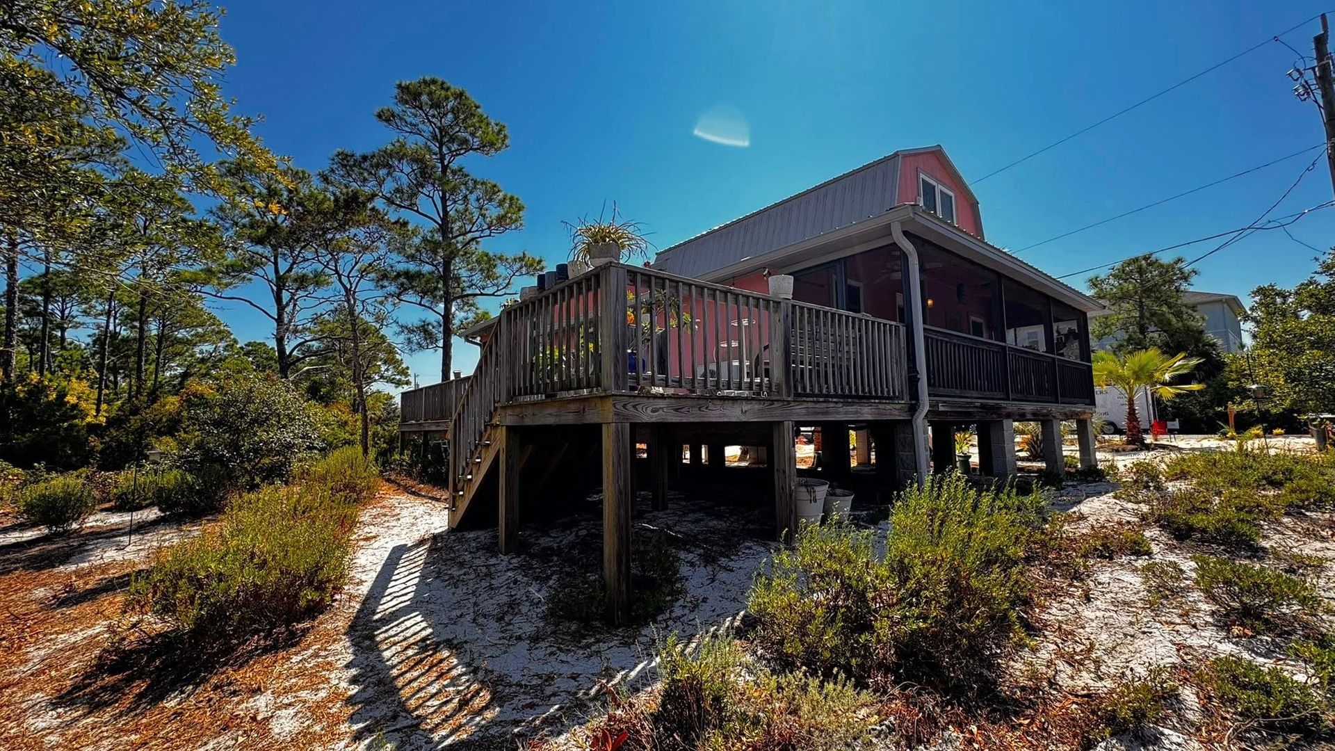 A wooden house on stilts sits in a sandy, tree-filled landscape under a clear blue sky.